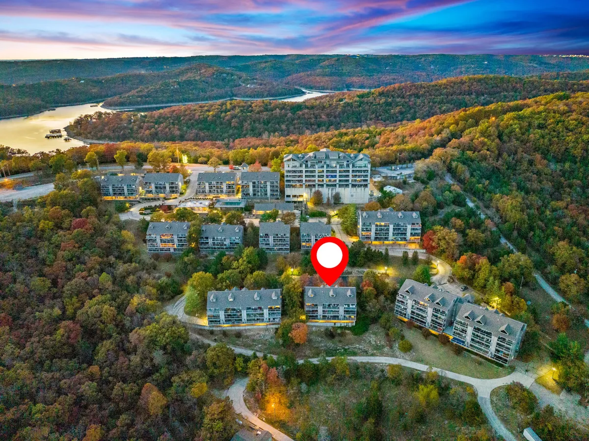 A picturesque exterior shot showing the modern condo buildings nestled among colorful autumn foliage, with a sweeping view of the lake in the background. The setting sun casts a warm glow over the landscape, highlighting the natural beauty and inviting atmosphere of the area.