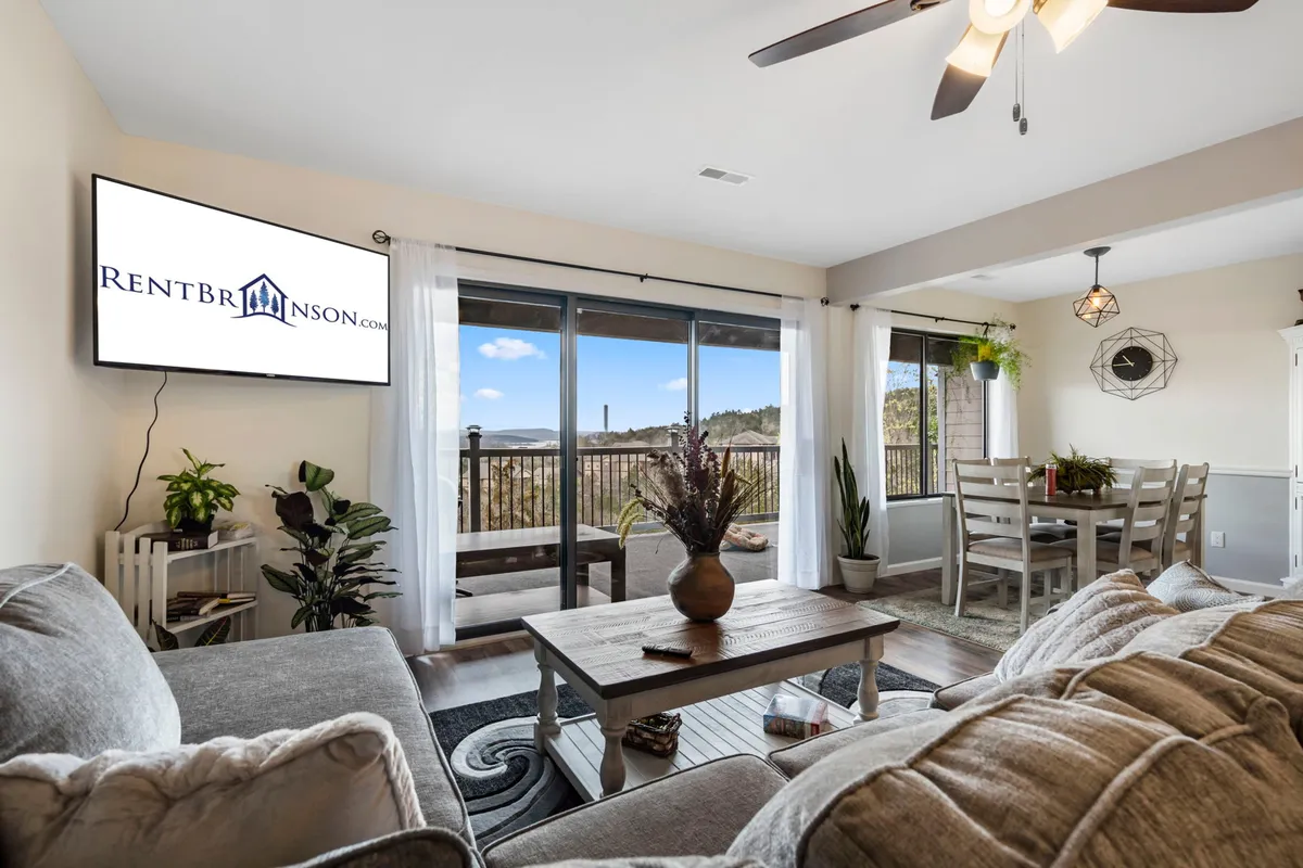 The image features a bright, modern living area with a plush sofa set beside expansive windows framing a stunning lake view. A stylish dining table invites shared meals, while a large TV promises cozy nights in—all set within an airy, modern farmhouse design.