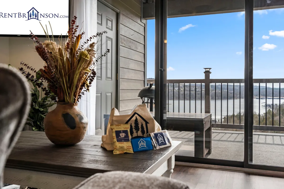 An interior scene focusing on a wooden coffee table with a decorative vase, set before glass doors that offer a picturesque view of water outside. The mood is cozy and tranquil, with sunlight streaming in, and the outdoors very much present through large windows.