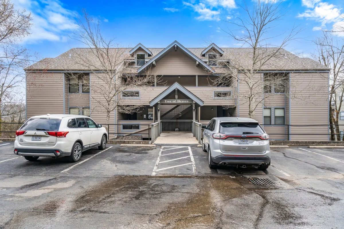 An exterior view of the property’s building entrance and parking area, framed by mature trees. The scene is welcoming, with easy-to-spot parking spaces close to the entrance, subtly suggesting convenience for guests with boats or trailers.
