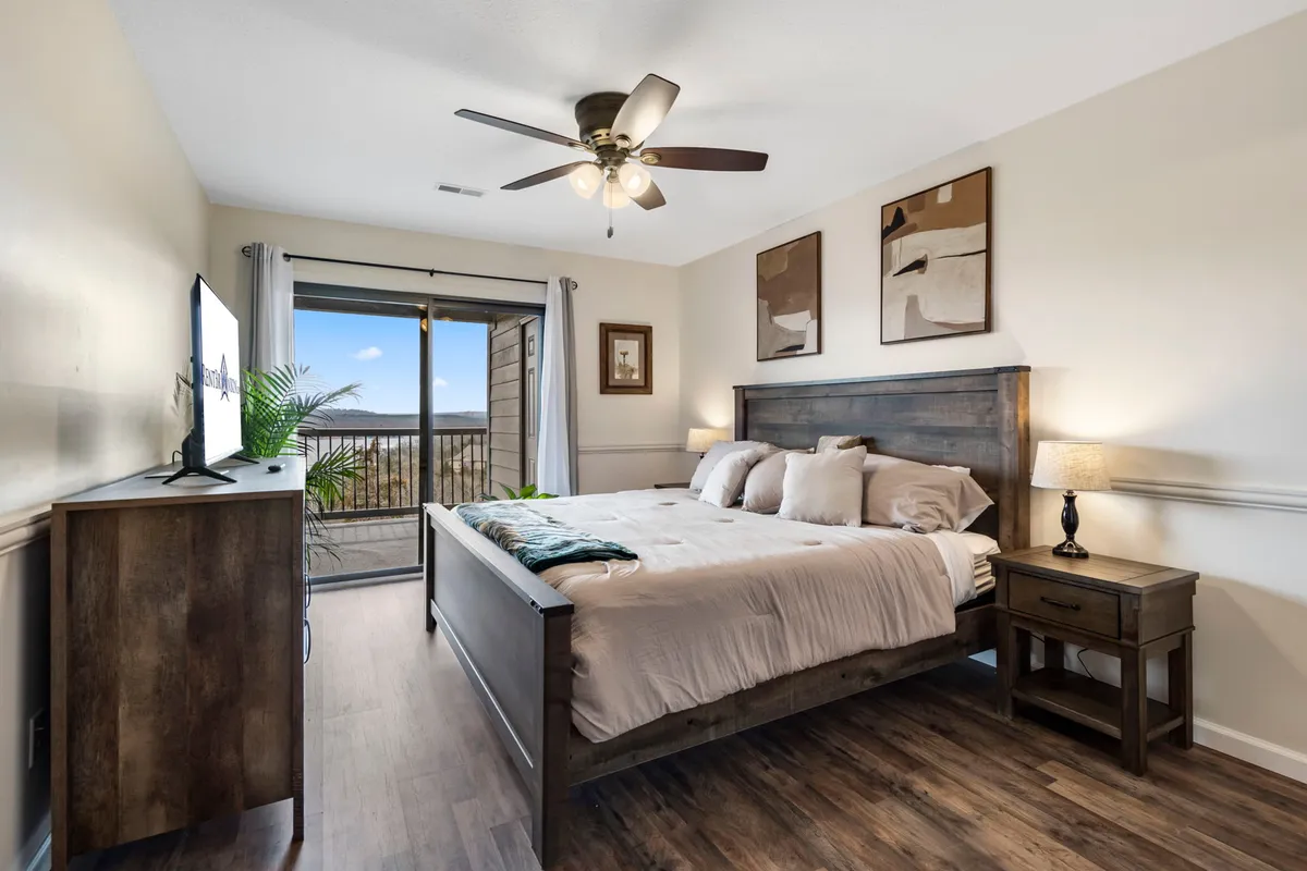 A bright bedroom featuring a bed with decorative pillows, a large window letting in natural light, a nightstand with a lamp, and a television. The scene feels serene and welcoming, ideal for restful nights or quiet mornings.