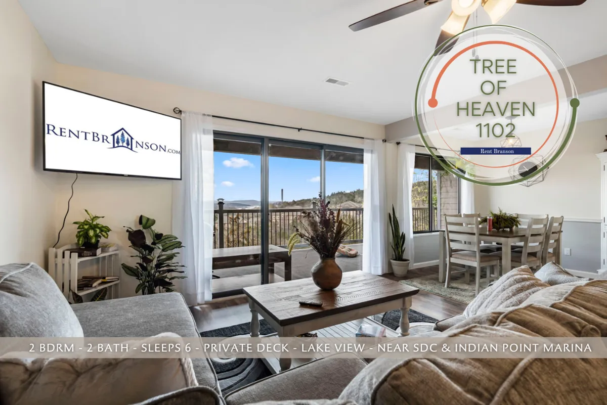 This image features a spacious, light-filled living area with a large window offering a sweeping view of Table Rock Lake. The scene includes a comfortable couch, a wooden dining table set for meals, and a flat-screen TV. Natural light floods the space, and the open layout connects lounging and dining areas, with the lake view always in sight.