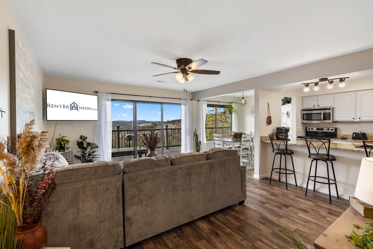 The image presents a cohesive view of the open living, dining, and kitchen area. Windows along the wall offer glimpses of the outdoors, while the space is filled with inviting furniture, a dining table, and a kitchen area for gathering and meals. The overall mood is comfortable and welcoming, with farmhouse-chic touches visible.